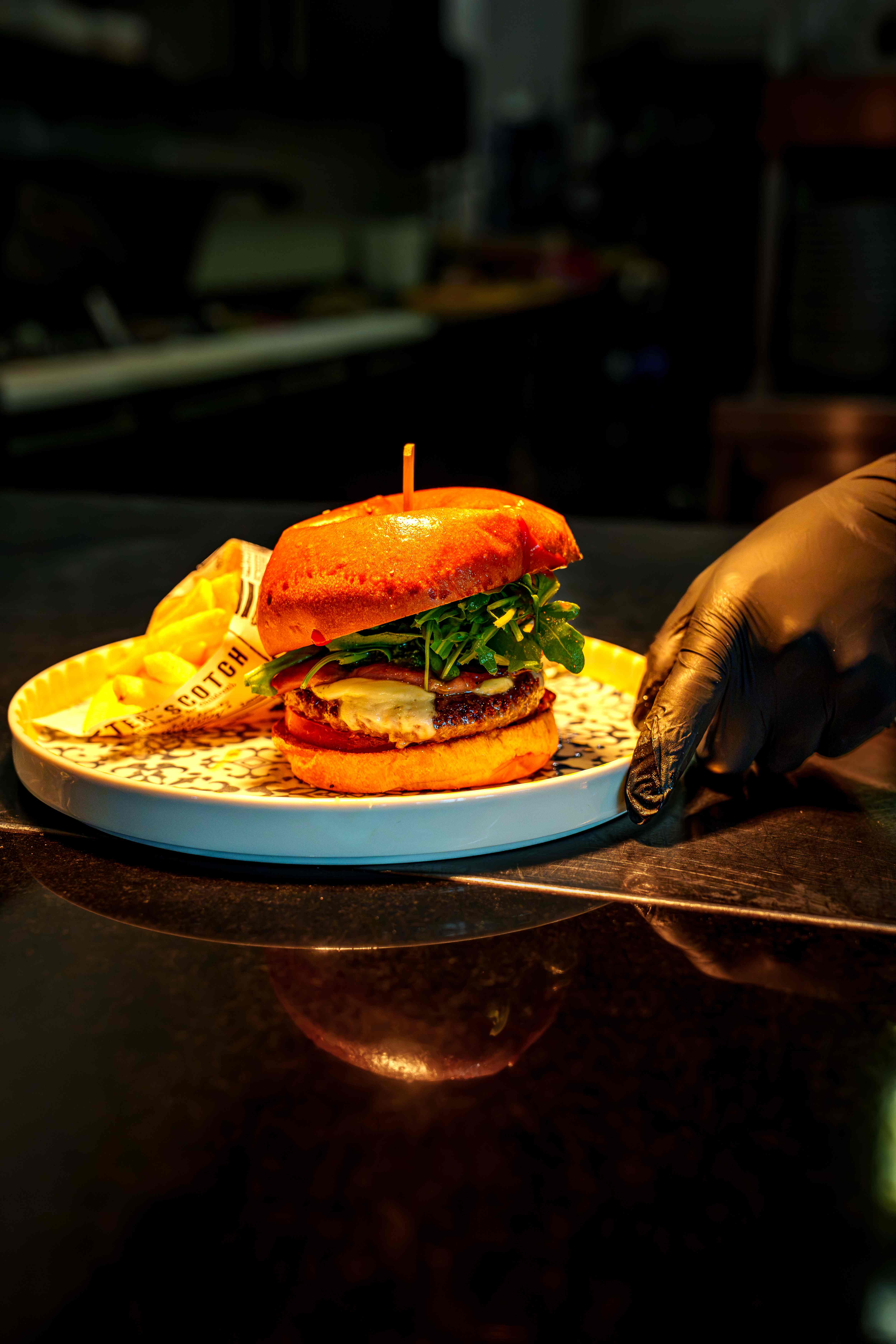 Burger im Brioche-Brötchen mit Käse und Rucola, dazu Pommes in der Papiertüte auf einem gemusterten Teller