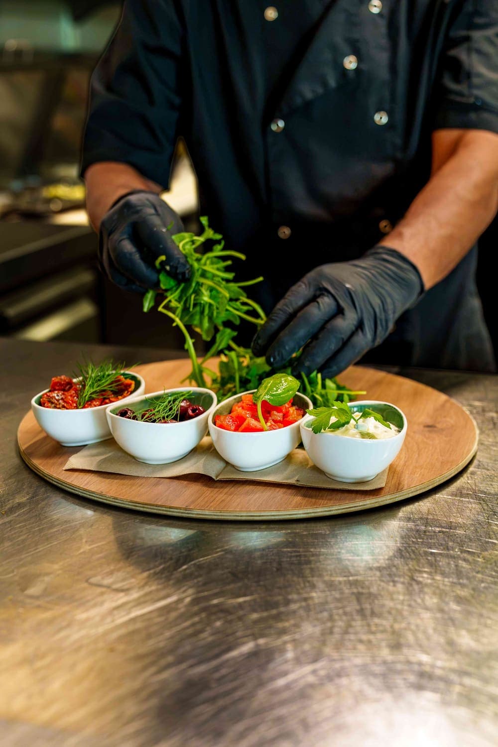 Koch mit schwarzen Handschuhen richtet Rucola und kleine Schalen mit Tomaten, Oliven und Dip an