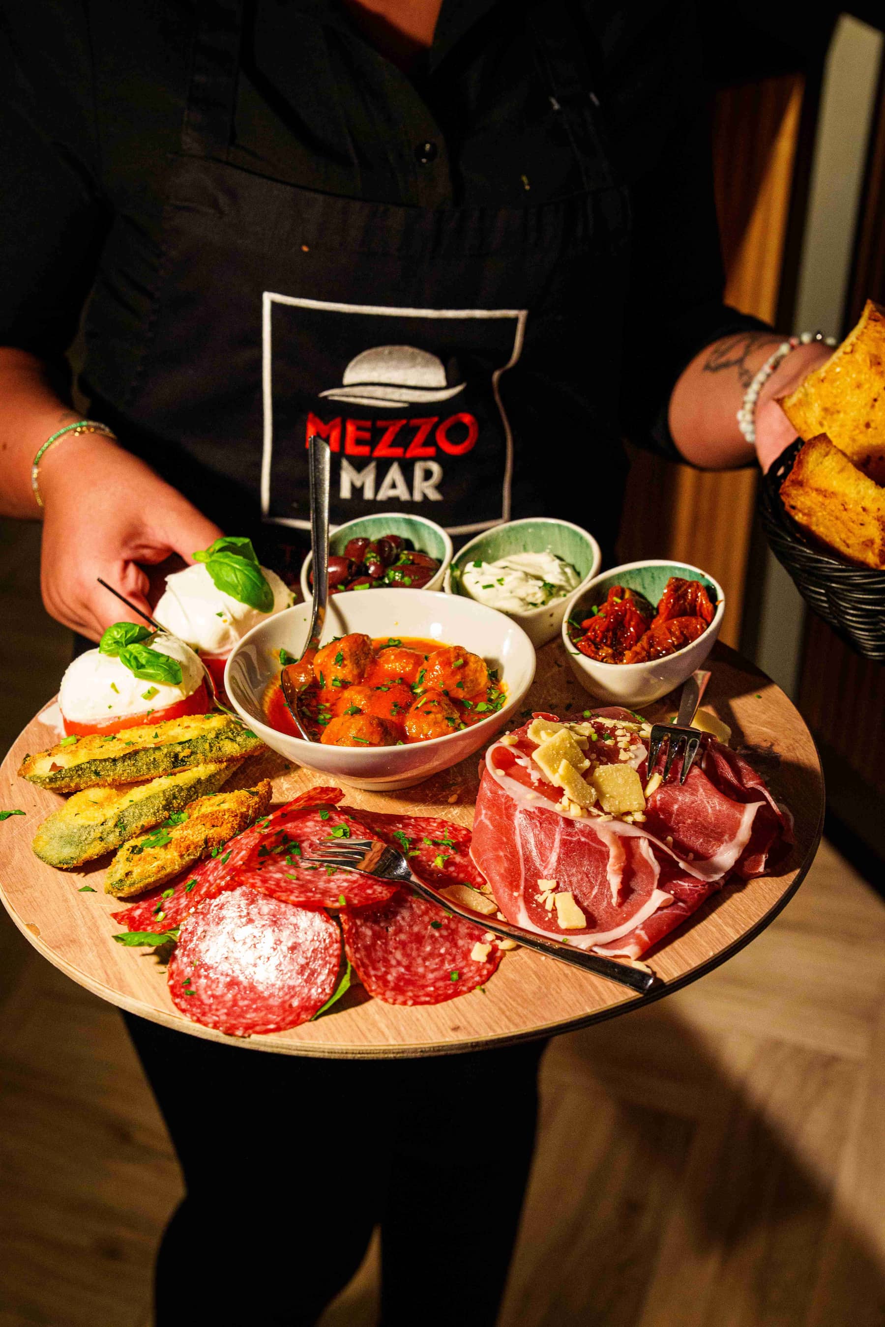 Servicekraft mit Mezzomar-Schürze serviert eine bunte Antipasti-Platte mit Mozzarella, Fleischbällchen, Salami und Schinken