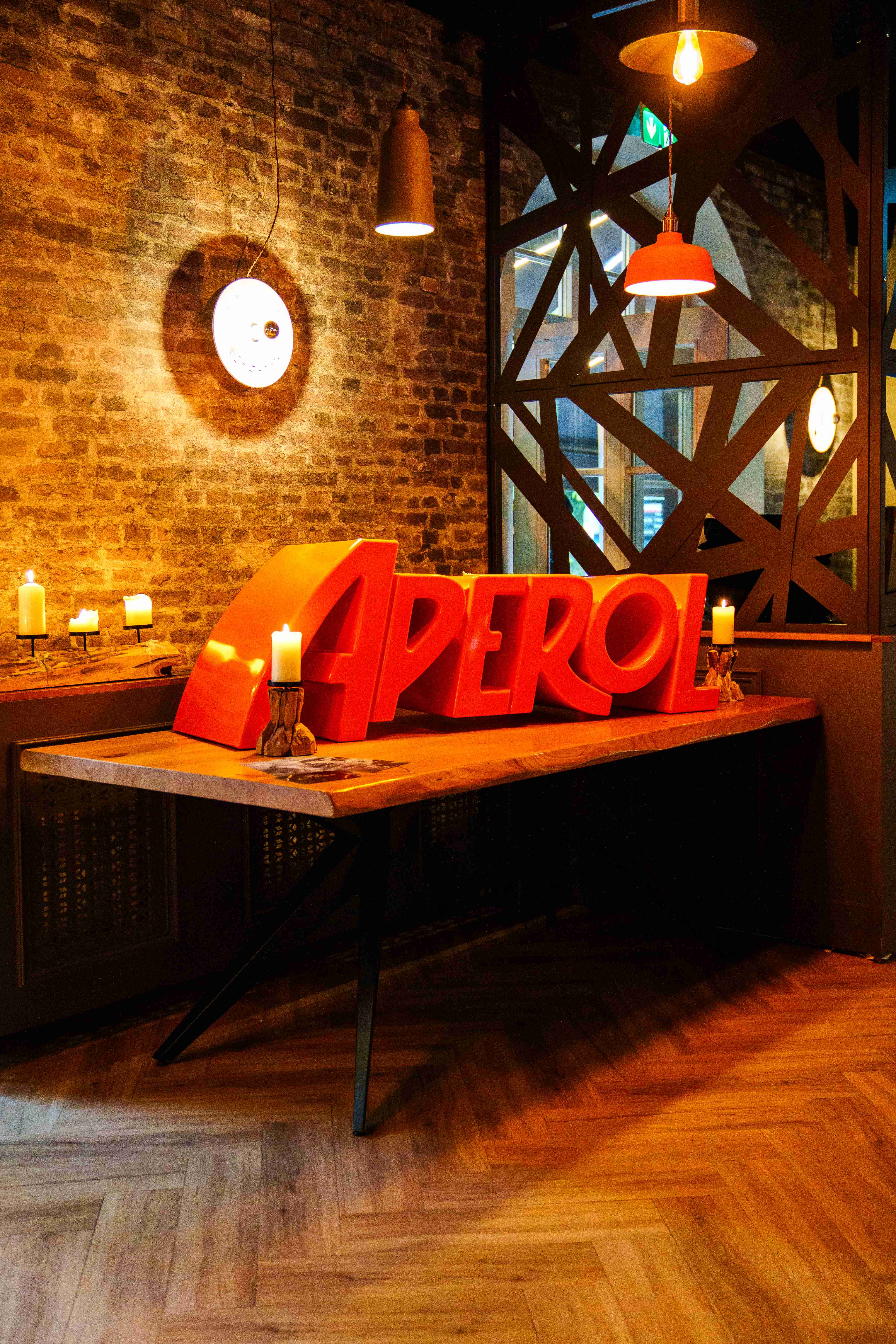 Leuchtender Aperol-Schriftzug auf einem Holztisch mit Kerzen vor einer Backsteinwand im Mezzomar