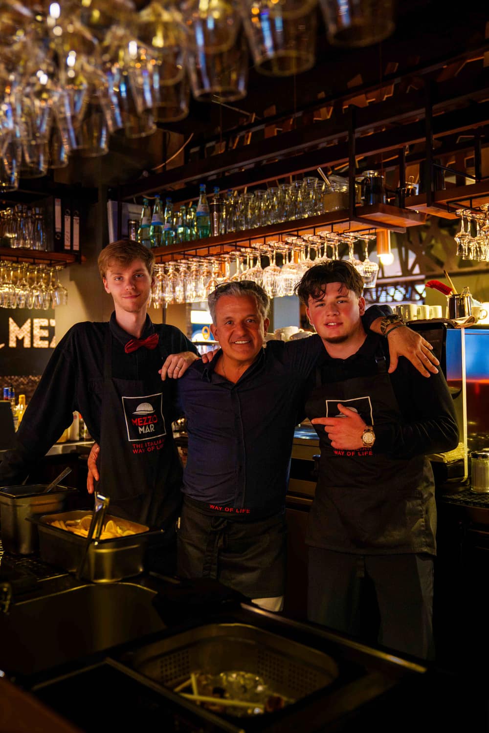 Drei Mitarbeiter des Mezzomar mit Mezzomar-Schürzen lächelnd hinter der Bar mit hängenden Gläsern