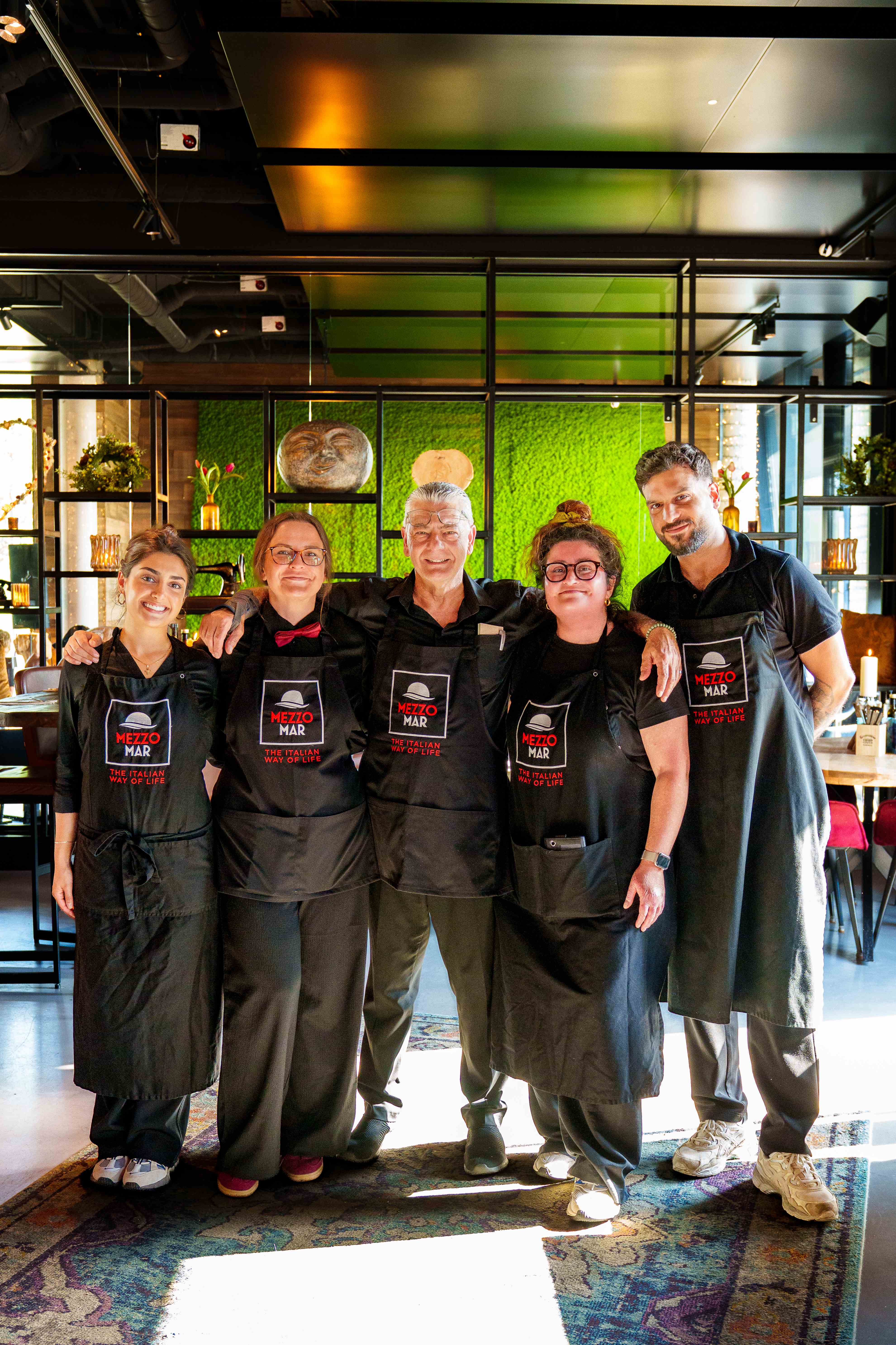 Das Mezzomar-Team in schwarzen Schürzen steht lächelnd vor der grünen Moosgras-Wand im Restaurant